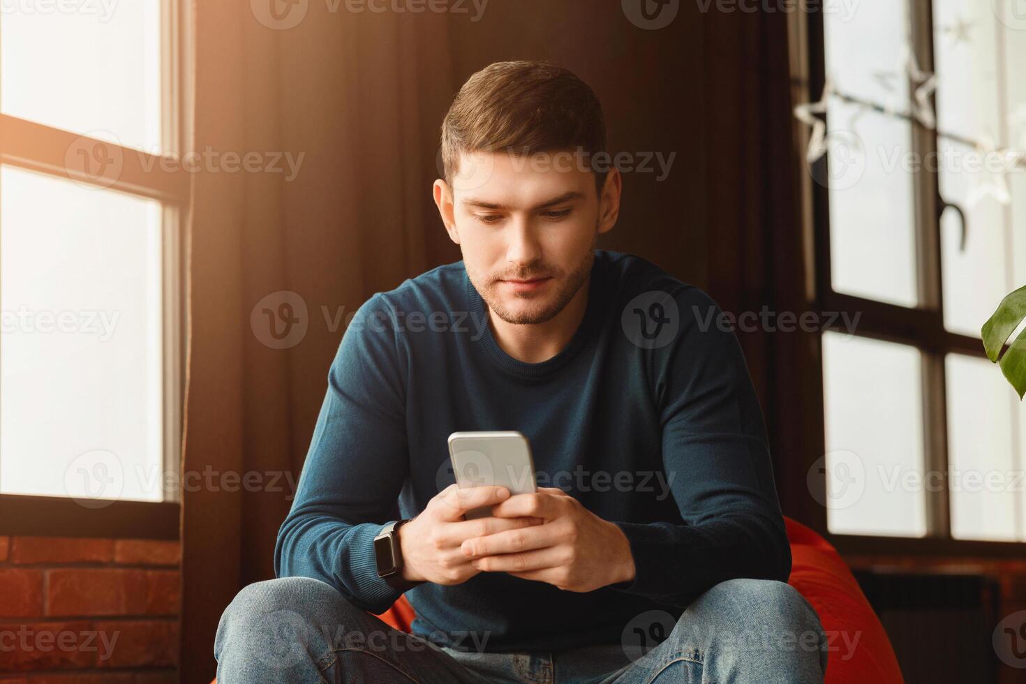 Millennial Guy Using Smartphone Texting Sitting In Beanbag Chair In Modern Loft Interior Indoor. Selective Focus photo