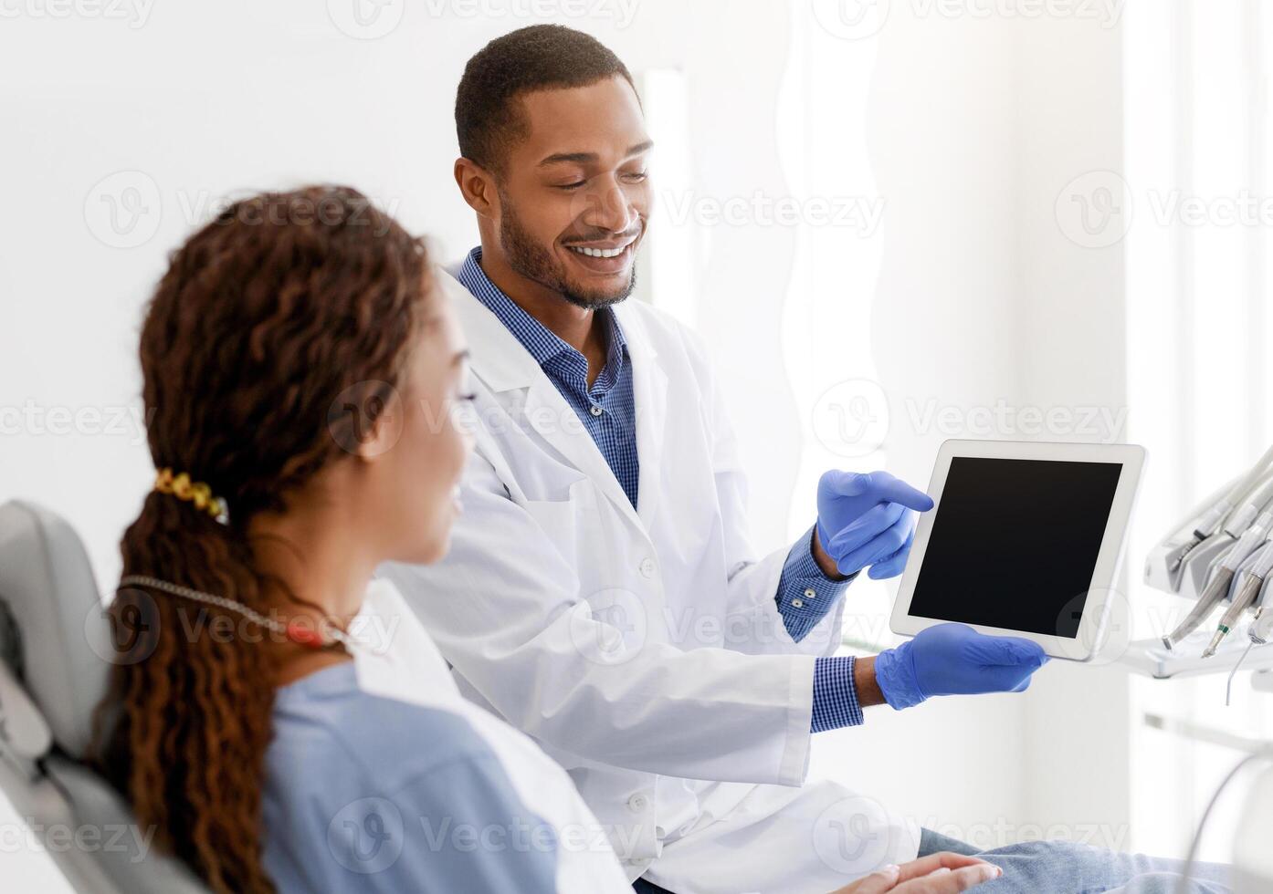Handsome smiling man dentist showing female patient empty digital tablet screen, clinic interior, copy space photo