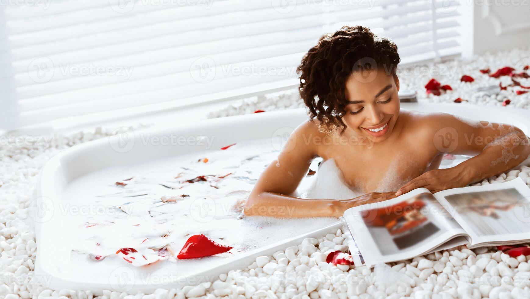 Relaxing day. Happy black lady taking bath with rose peals and reading magazine at home photo