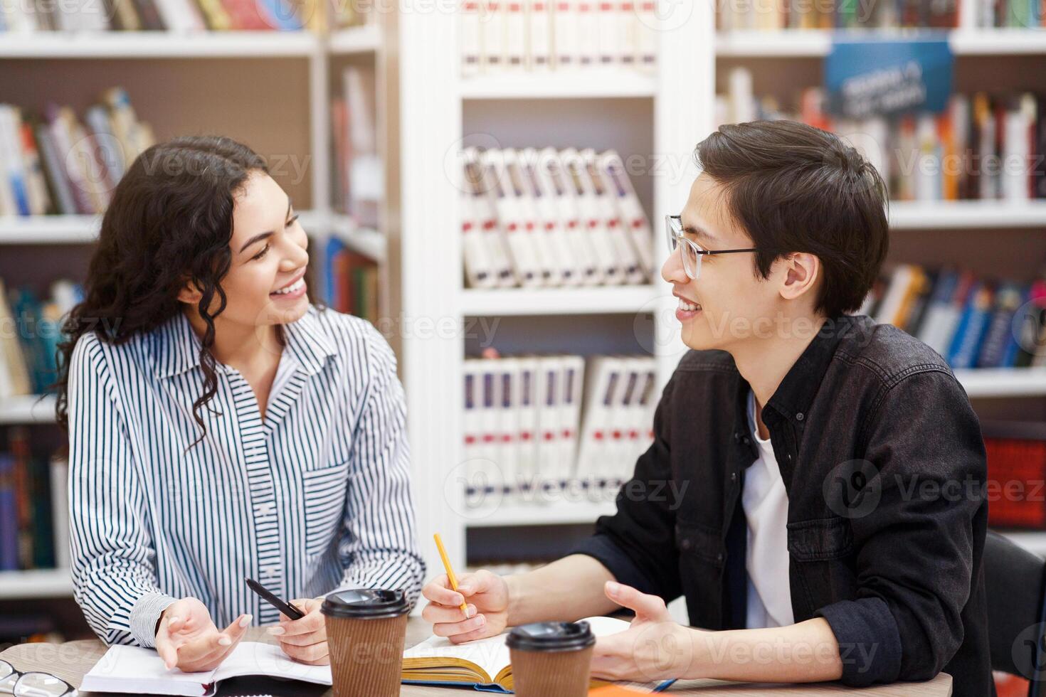 Two happy multiracial students talking at looking at each other sitting at desk in campus library, doing homework photo