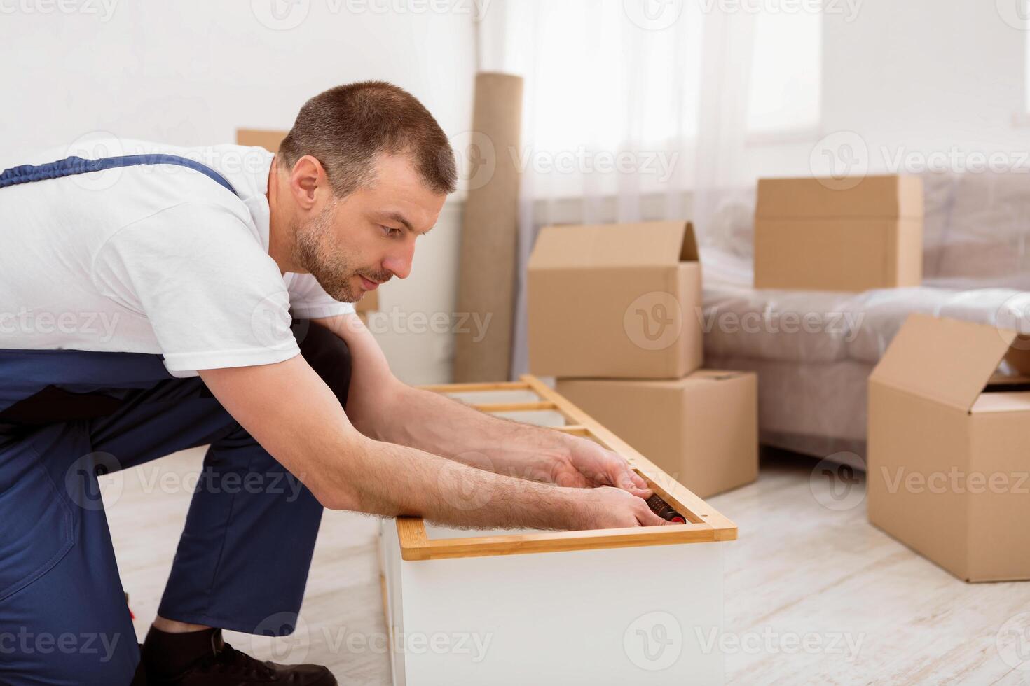 Repairman In Blue Coverall Fixing And Assembling Wooden Shelf With Screwdriver During Renovation In Flat Indoors. Furniture Assembly And Carpentry Service Concept. Side View Shot photo