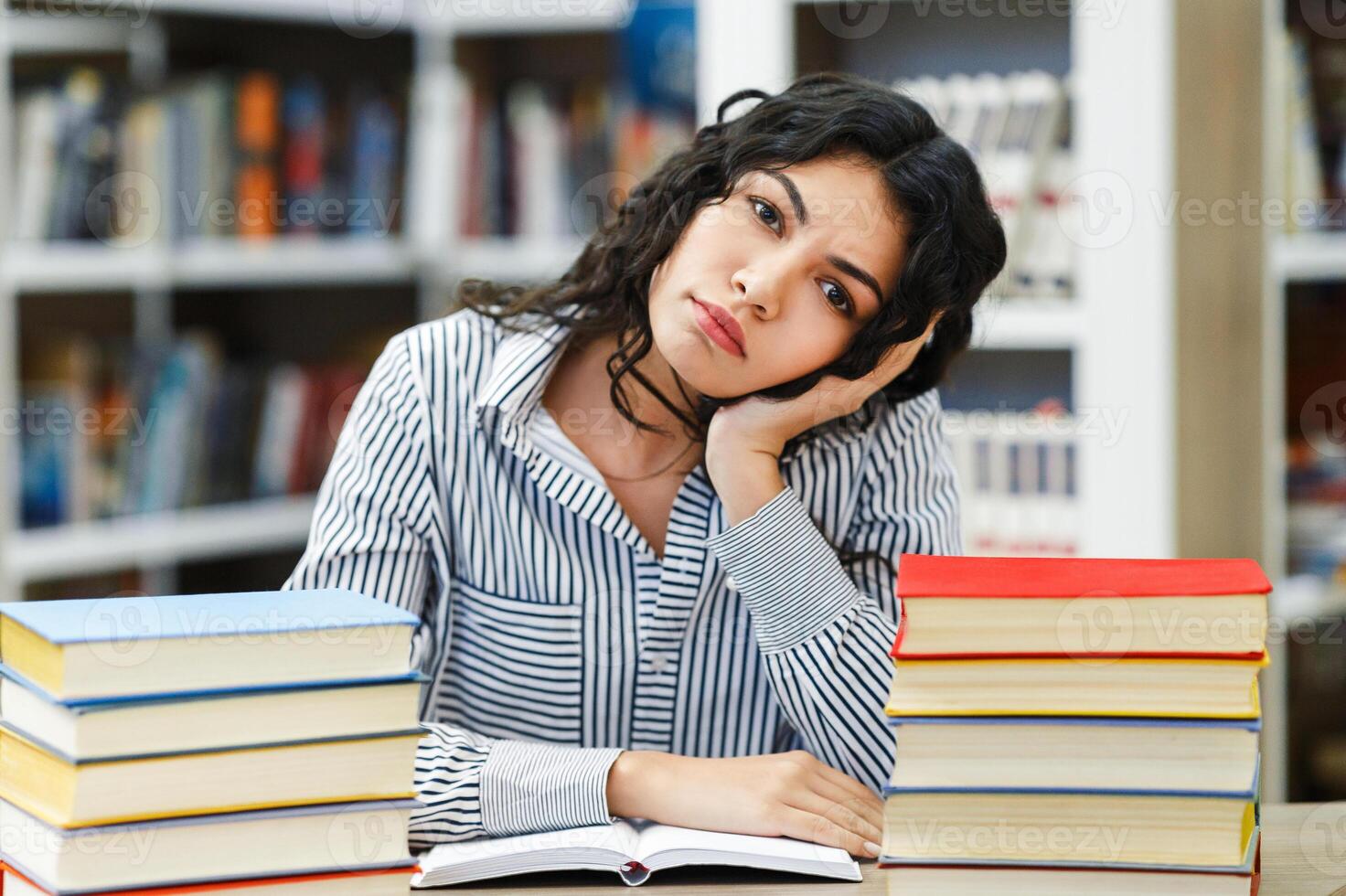 Boring Homework Concept. Tired latin female student sitting at desk with many books in public library, copyspace photo