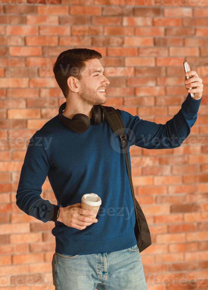 College Student Making Selfie On Smartphone Holding Paper Coffee Cup Posing On Brick Wall Indoor. Youth Culture And Modern Lifestyle photo