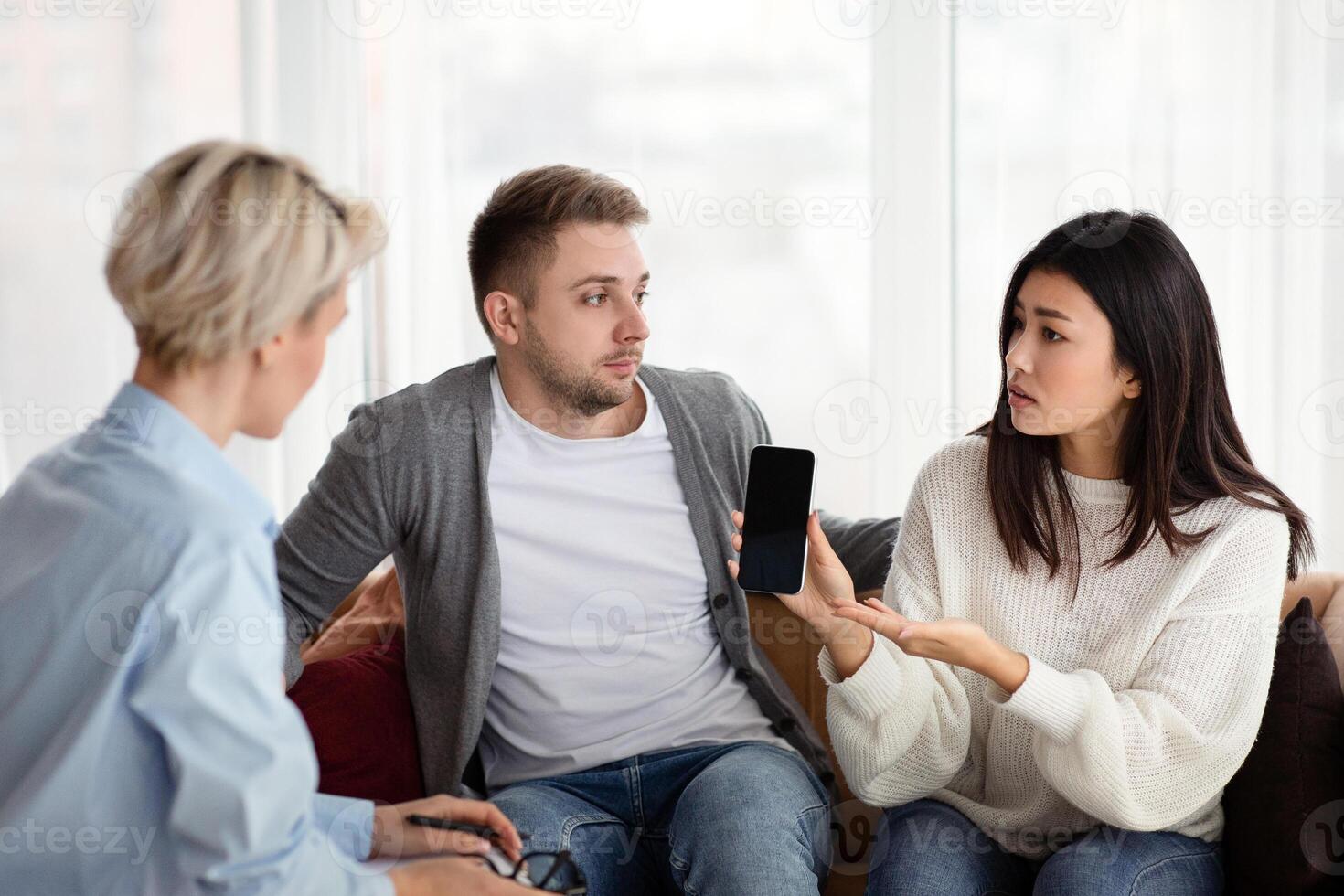 Wife Showing Cheating Husband's Cellphone Blaming Him During Family Therapy Session With Psychologist In Office. Selective Focus photo