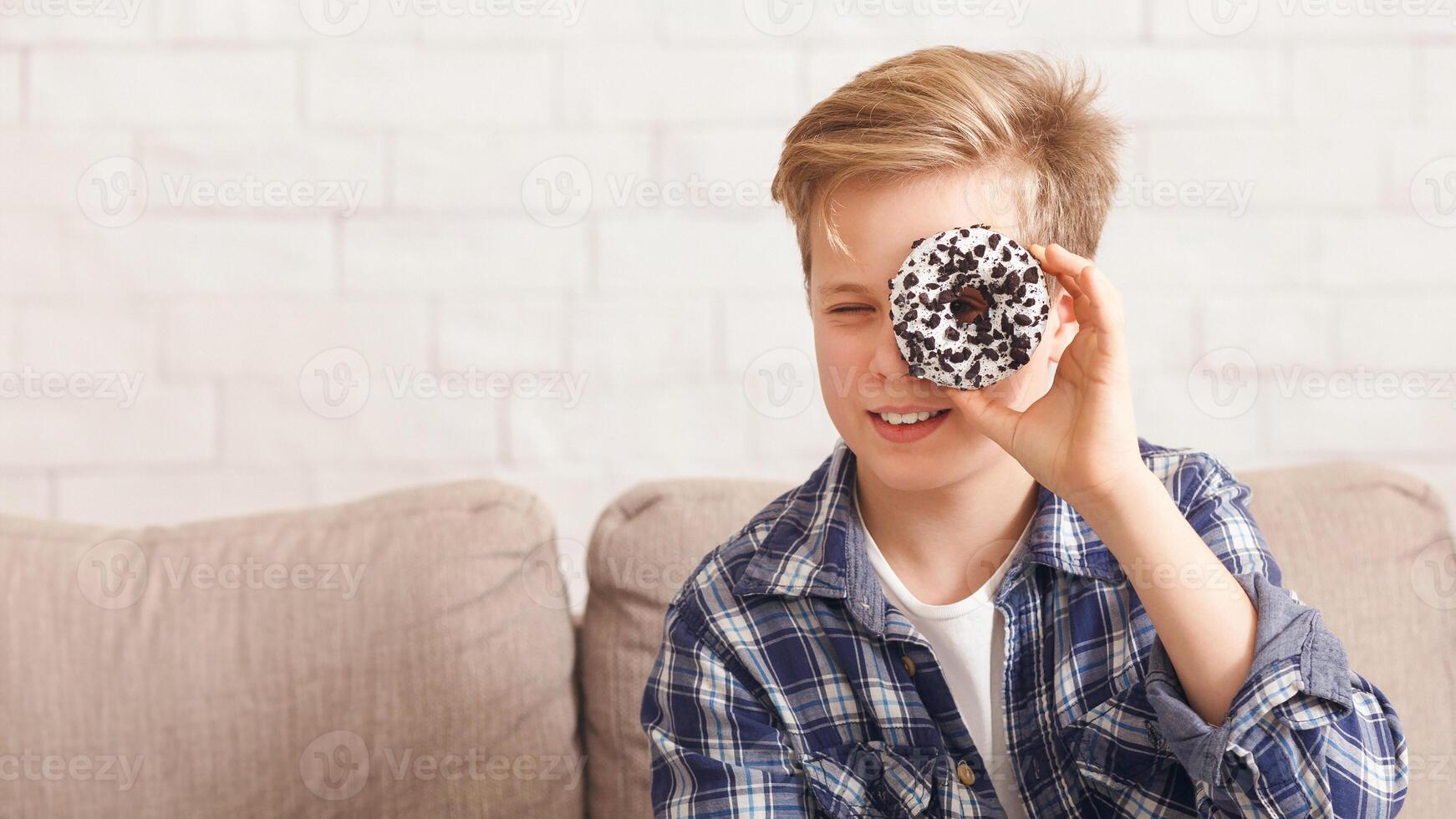 Boy Holding Doughnut Near Eye Posing Having Fun With Junk Food Sitting On Couch At Home. Panorama, Copy Space photo