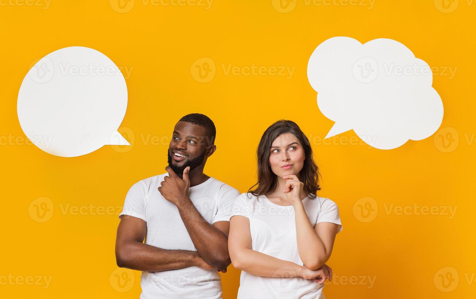 Portrait of thoughtful interracial couple looking at empty speech bubbles above their heads and touching chin photo
