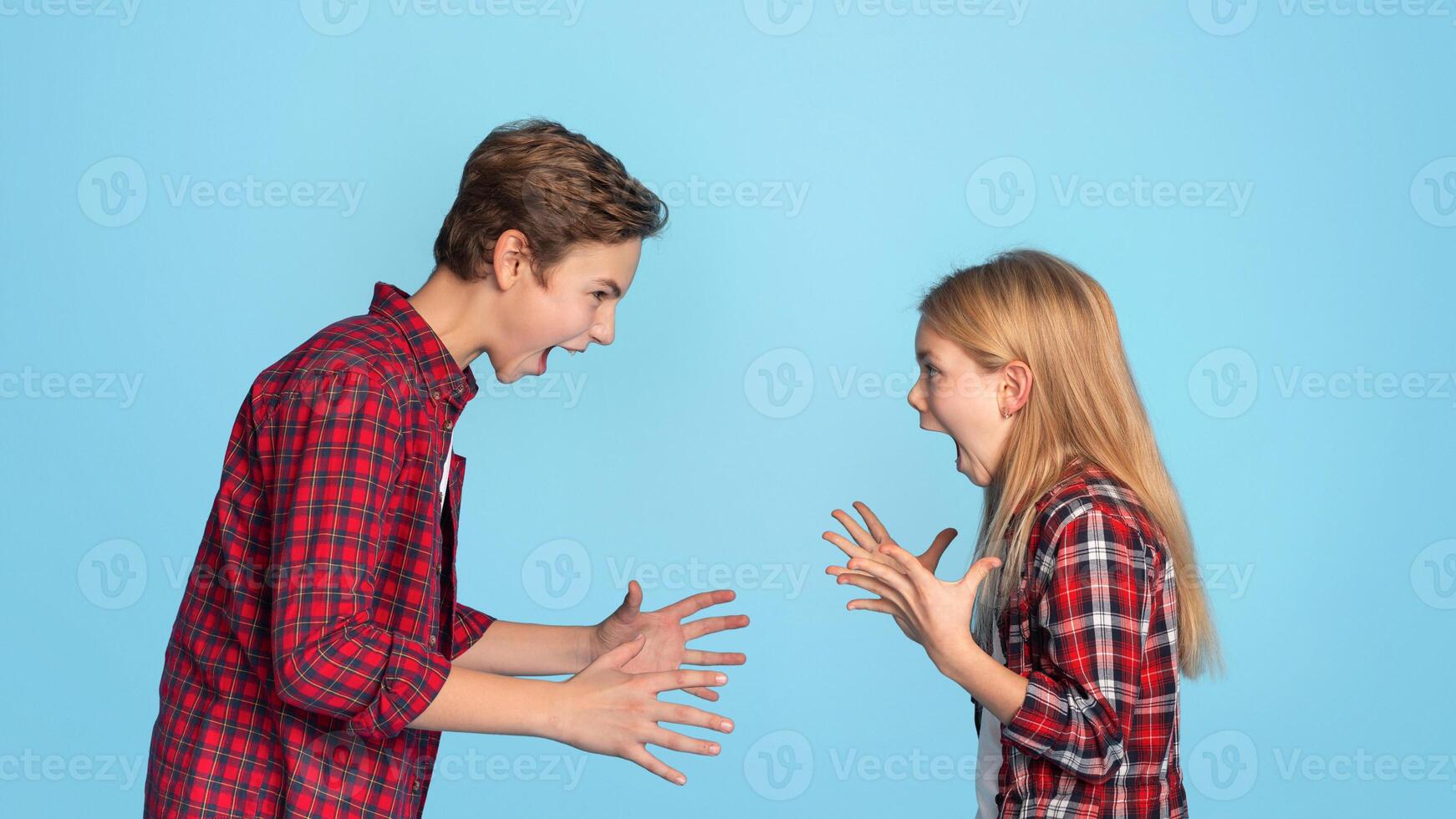 Siblings Conflicts. Angry Little Brother And Sister Emotionally Screaming At Each Other, Standing On Blue Background With Free Space photo