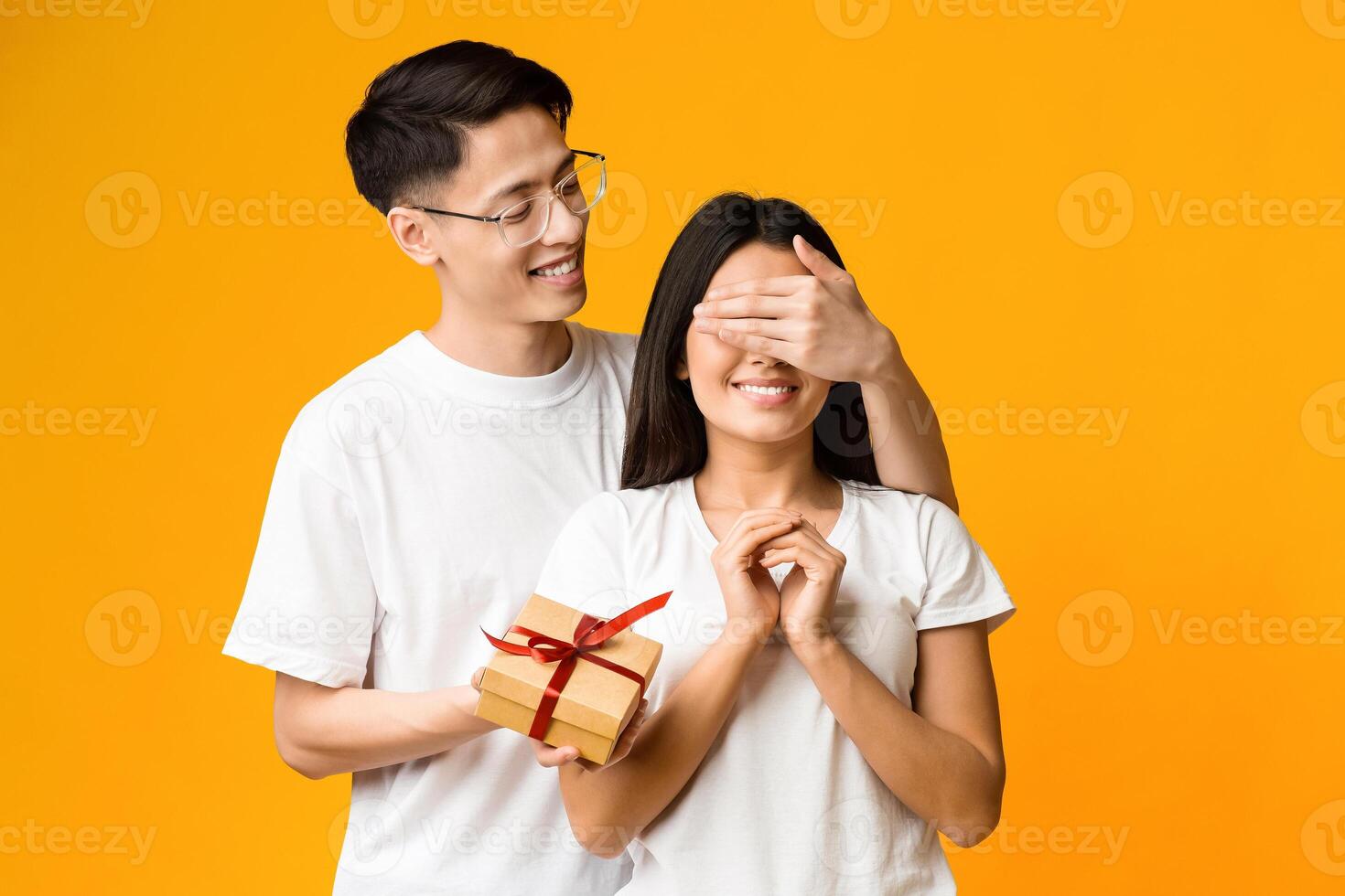 Cheerful asian guy making surprise to his excited girlfriend, covering her eyes, holding gift box photo