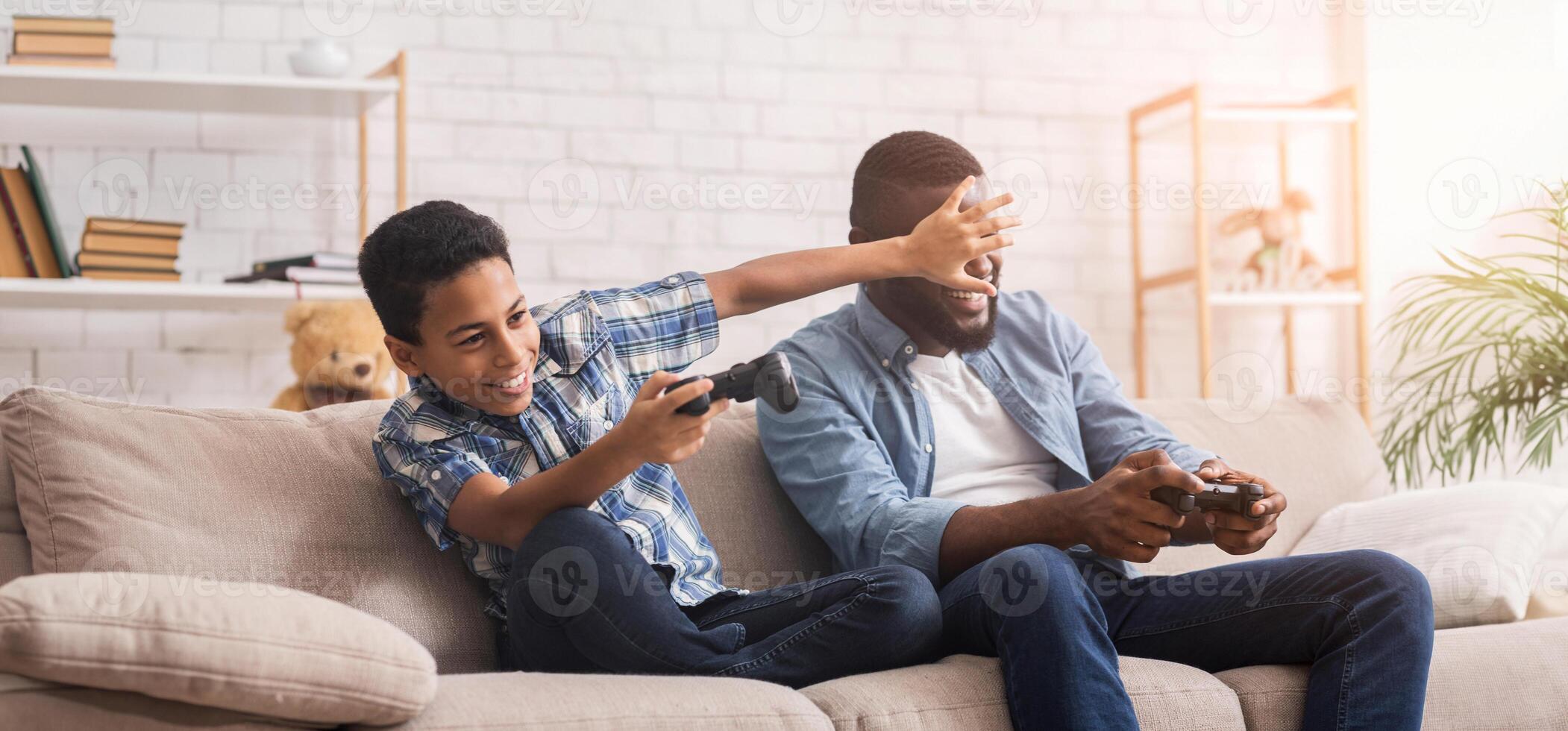 Cheerful afro dad and son with joysticks playing games at home, boy covering father's eyes with hand, free space photo