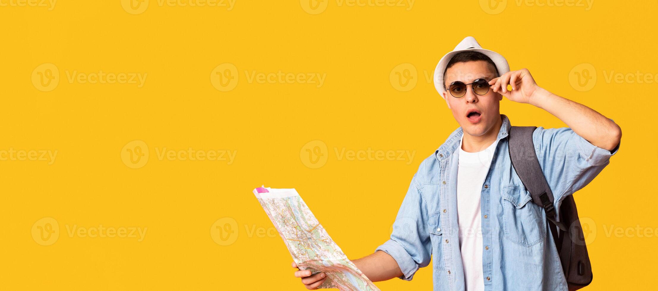 Amazed young backpacker with map, hat and sunglasses making sighseeing over orange background, panorama with copy space photo
