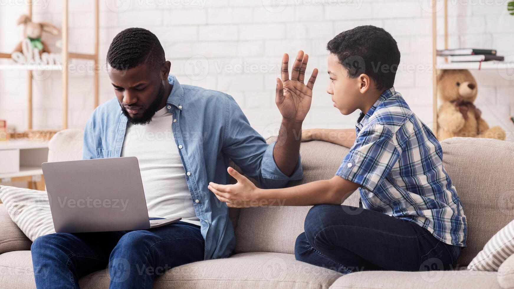 Little Afro Son Asking Attention From His Father Busy With Laptop, Working Man Ignoring His Kid And Showing Stop Gesture With Hand photo