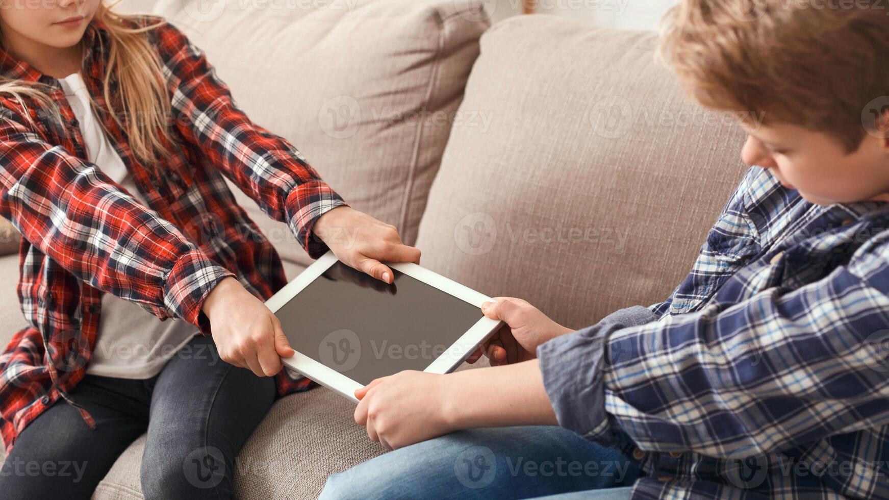 Jealous Brother And Sister Pulling Apart Tablet Not Wanting To Share Sitting On Sofa At Home. Siblings Relationship Problems. Cropped photo