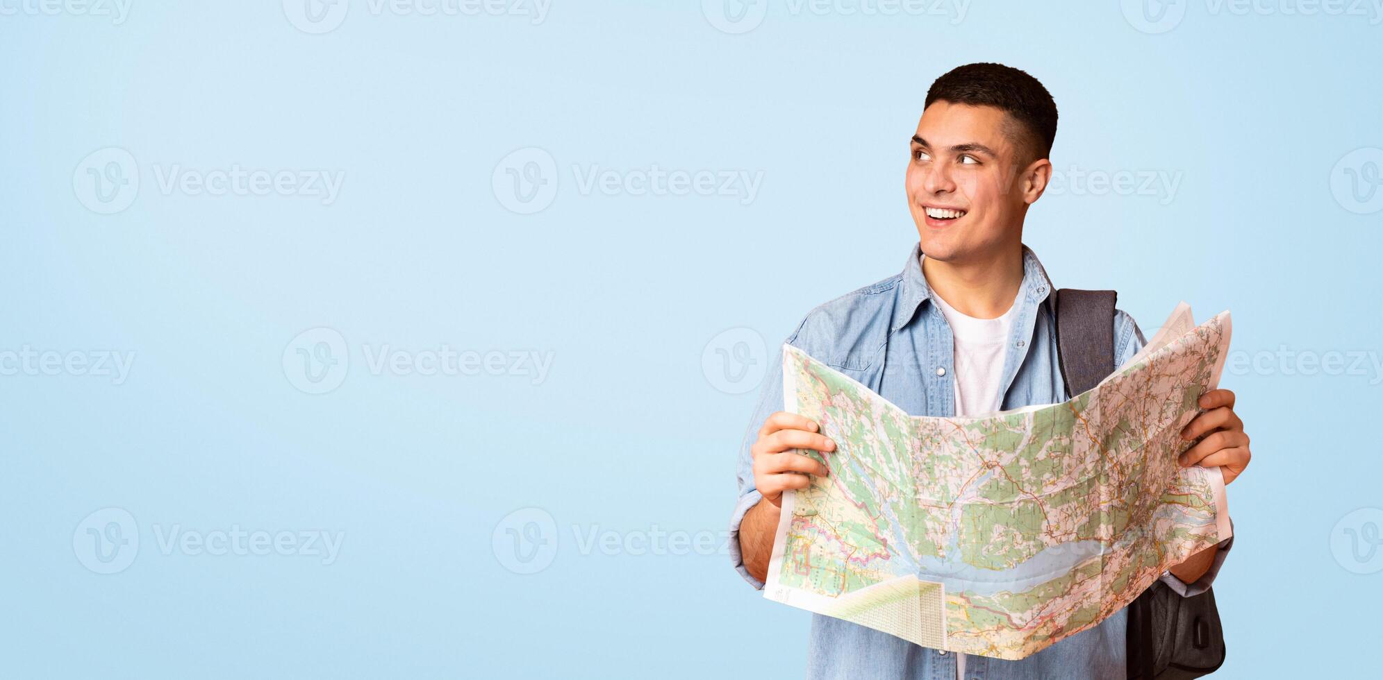 Happy young man traveler with backpack holding city map and looking at copy space, orange studio background, panorama photo