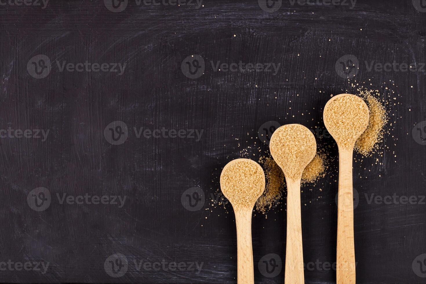 Brown cane sugar in three wooden spoons on black background. photo