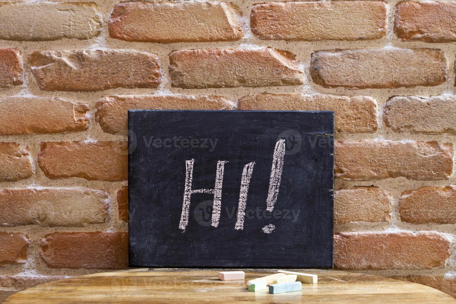 Black board with the word HI drown by hand on wooden table on brick wall background. photo