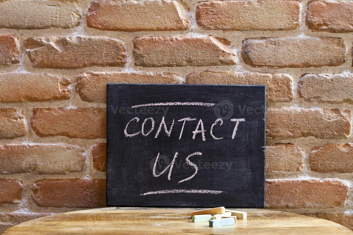 Black board with the phrase CONTACT US drown by hand on wooden table on brick wall background. photo