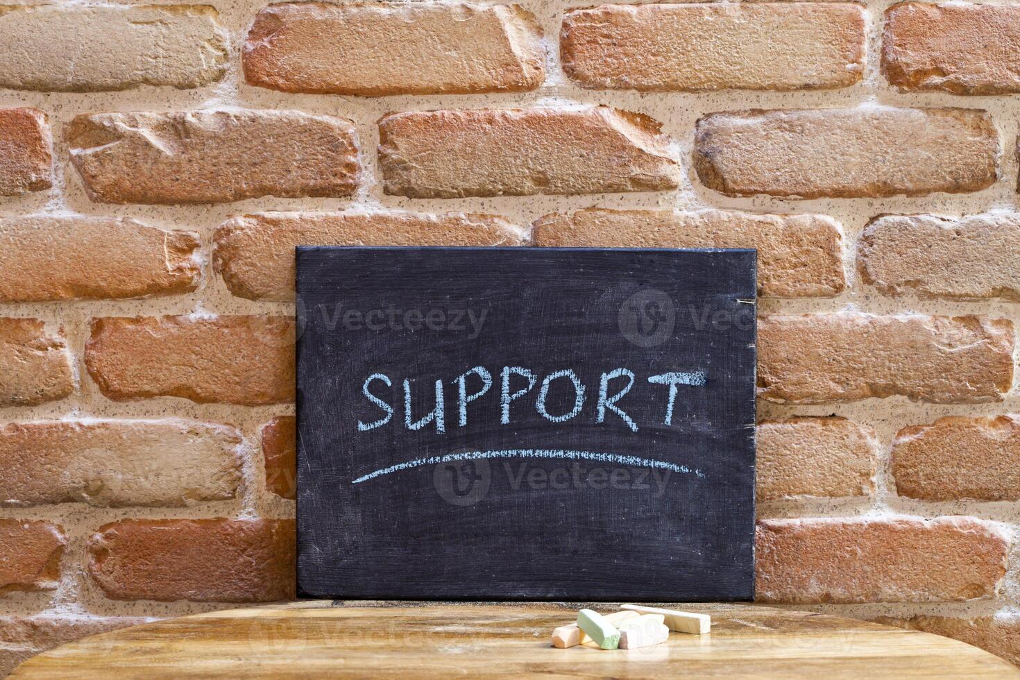 Black board with the word SUPPORT drown by hand on wooden table on brick wall background. photo