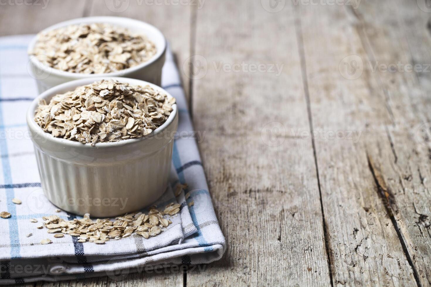 Oat flakes in ceramic bowls on linen napkin, golden wheat ears on rustic wooden background. photo