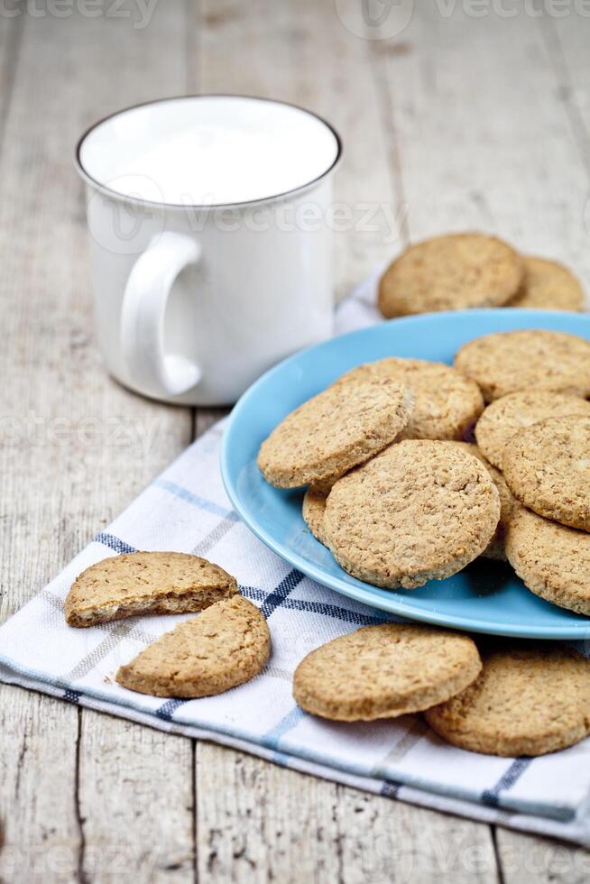 Fresh baked oat cookies on blue ceramic plate on linen napkin and cup of milk on rustic wooden table. photo