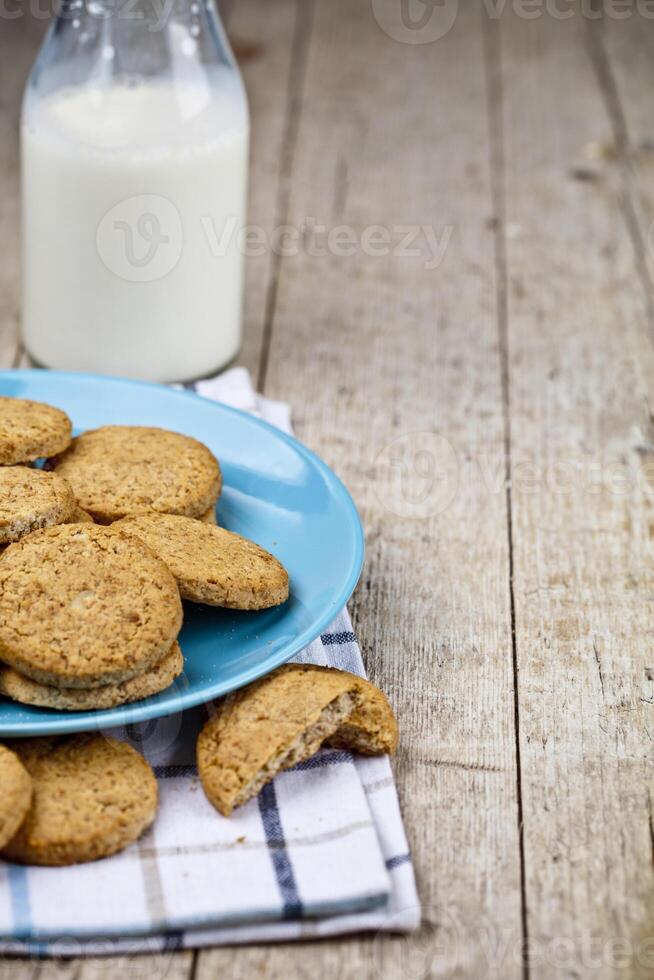 Fresh baked oat cookies on blue ceramic plate on linen napkin and bottle of milk on rustic wooden table background. photo