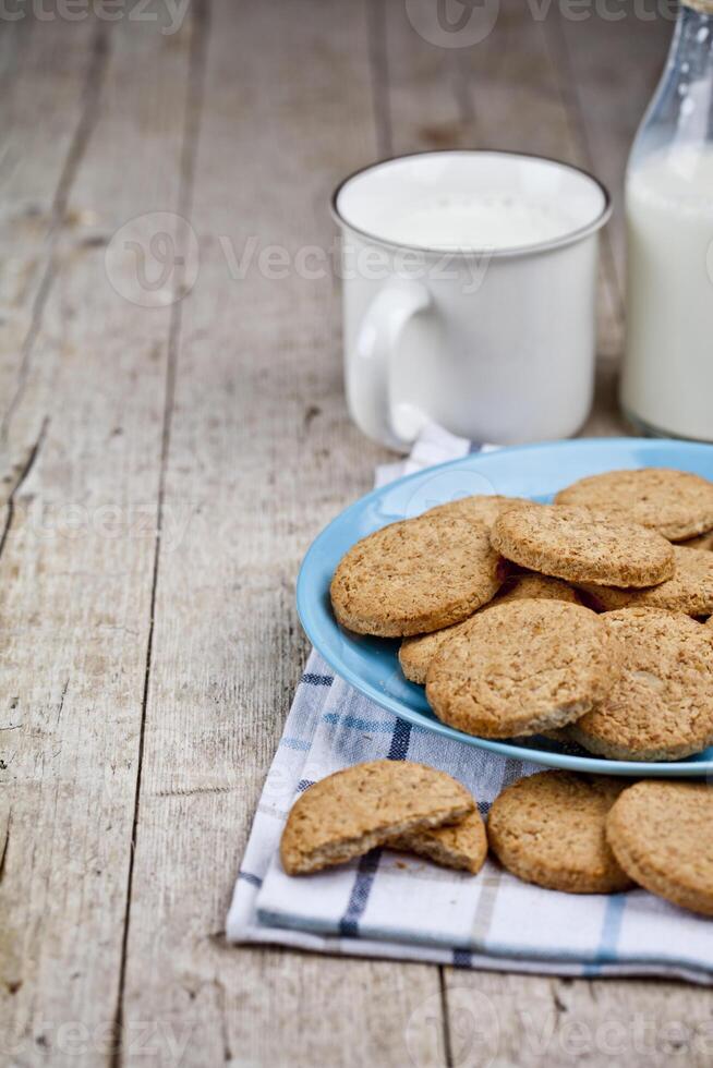 Fresh baked oat cookies on blue ceramic plate on linen napkin and cup of milk on rustic wooden table background. photo