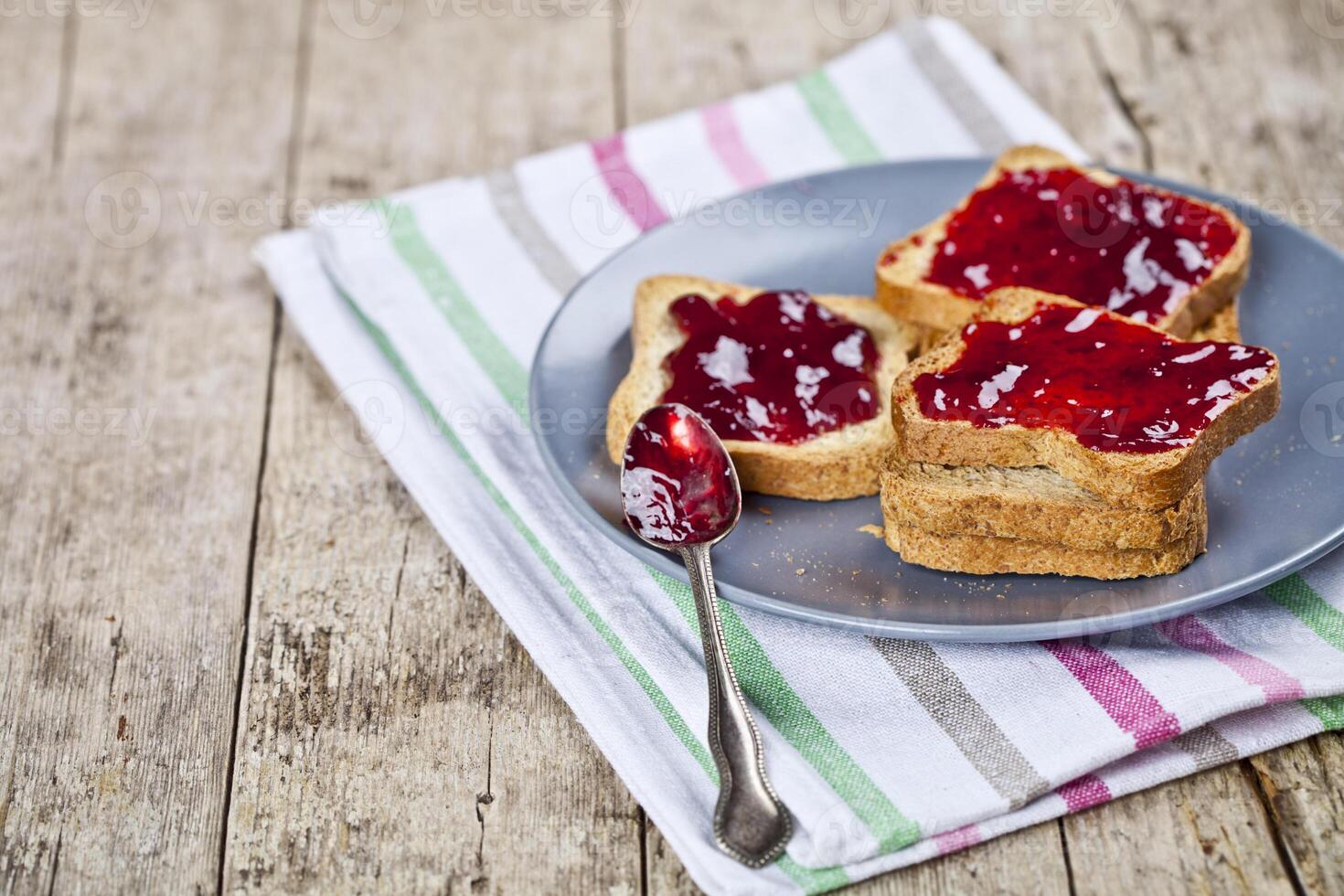 Fresh toasted cereal bread slices with homemade cherry jam and spoon on ceramic plate closeup on linen napkin on rustic wooden table background. photo