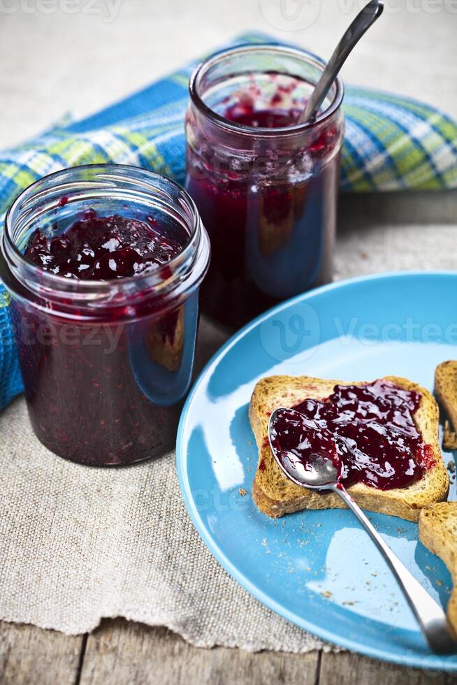 Fresh cereal bread slices on blue ceramic plate, homemade cherry and wild berries jam in jars and spoons closeup on linen napkin on rustic wooden table background. photo