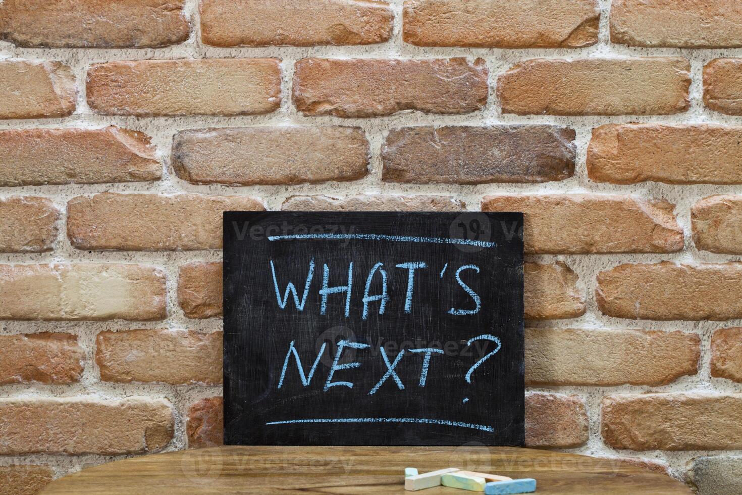 Chalk board with the phrase WHAT'S NEXT drown by hand and chalks on wooden table on brick wall background. photo