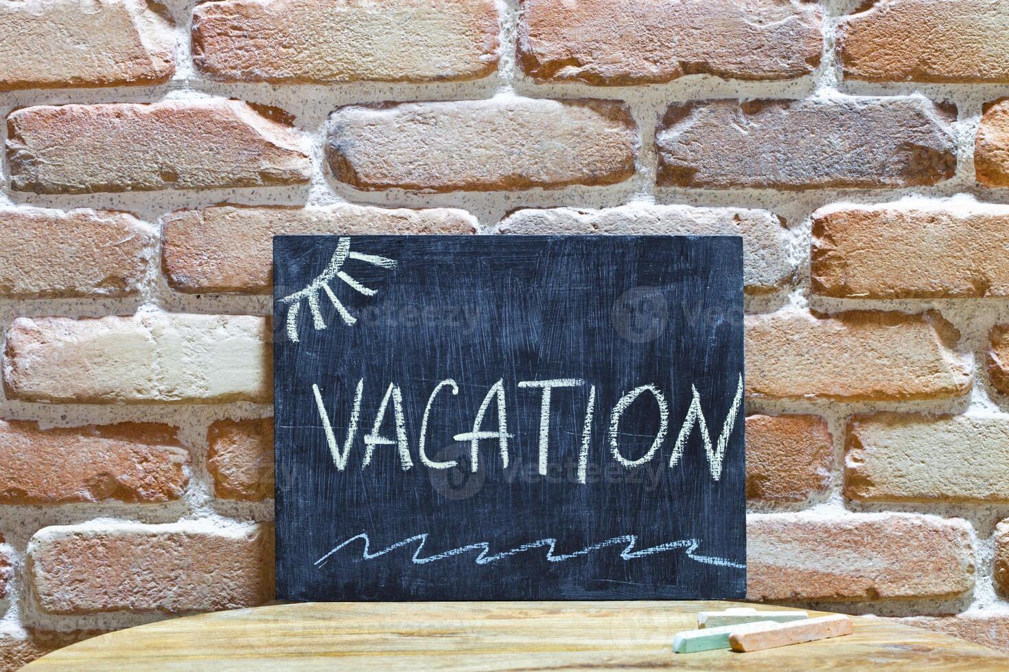 Chalk board with the word VACATION drown by hand and chalks on wooden table on brick wall background. photo