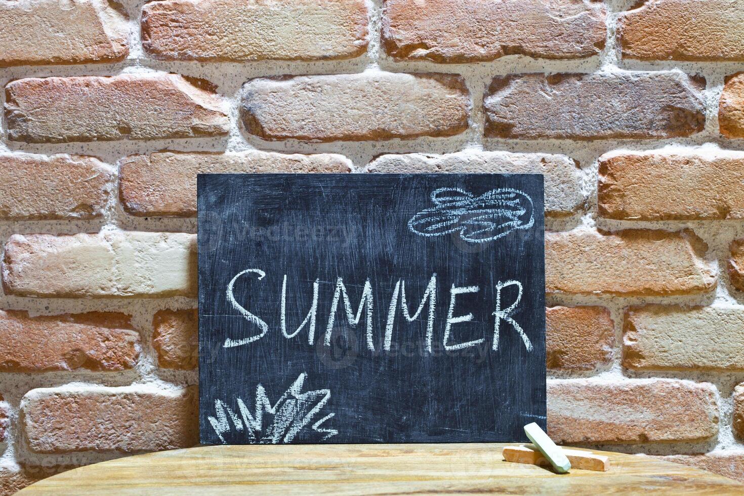 Chalk board with the word SUMMER drown by hand and chalks on wooden table on brick wall background. photo