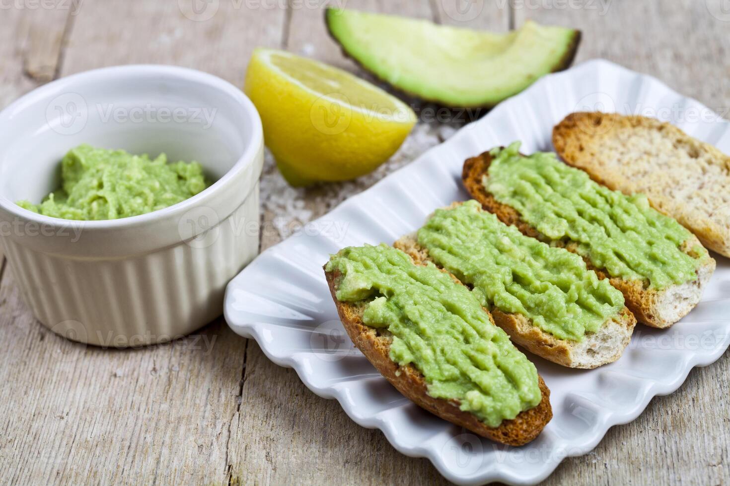 Crostini with avocado guacamole on white plate closeup on rustic wooden table. Diet breakfast. Delicious and healthy plant-based food. photo