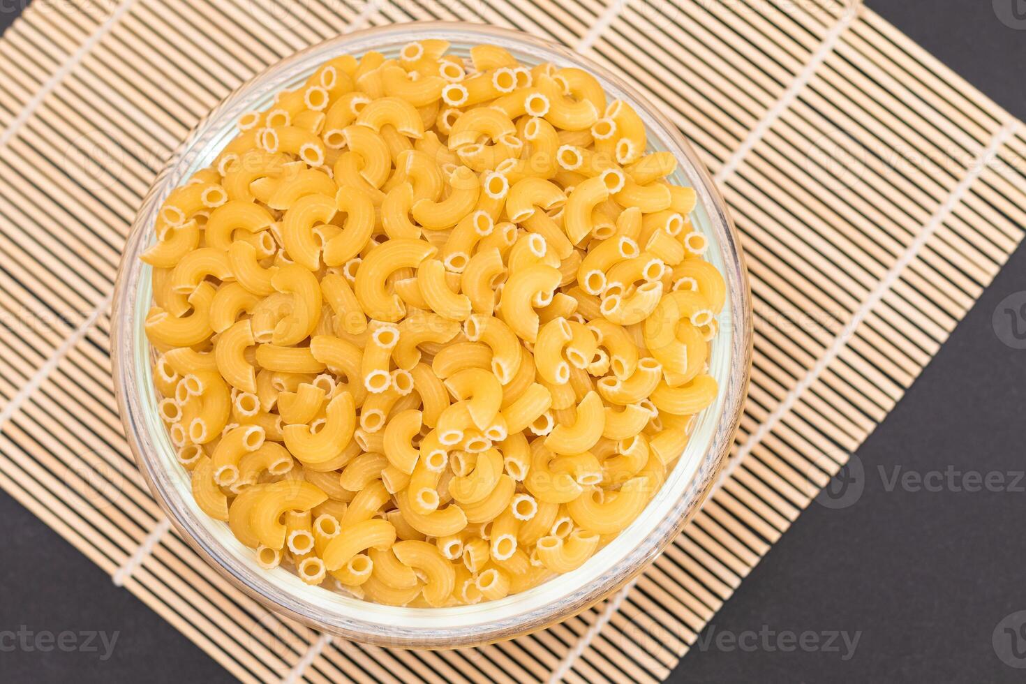 Uncooked Chifferi Rigati Pasta in Glass Jar on Bamboo Mat on Black Background. Fat and Unhealthy Food. Classic Dry Macaroni. Italian Culture and Cuisine. Raw Pasta photo
