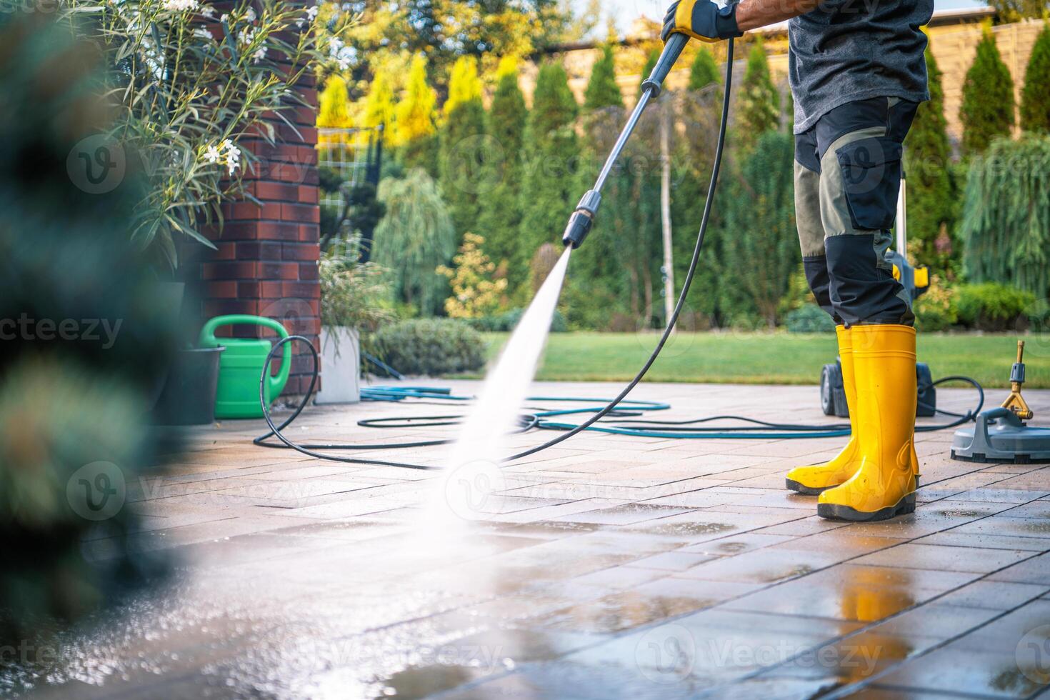 Person in Yellow Boots Pressure Washing a Patio Surrounded by a Lush Garden on a Sunny Day photo