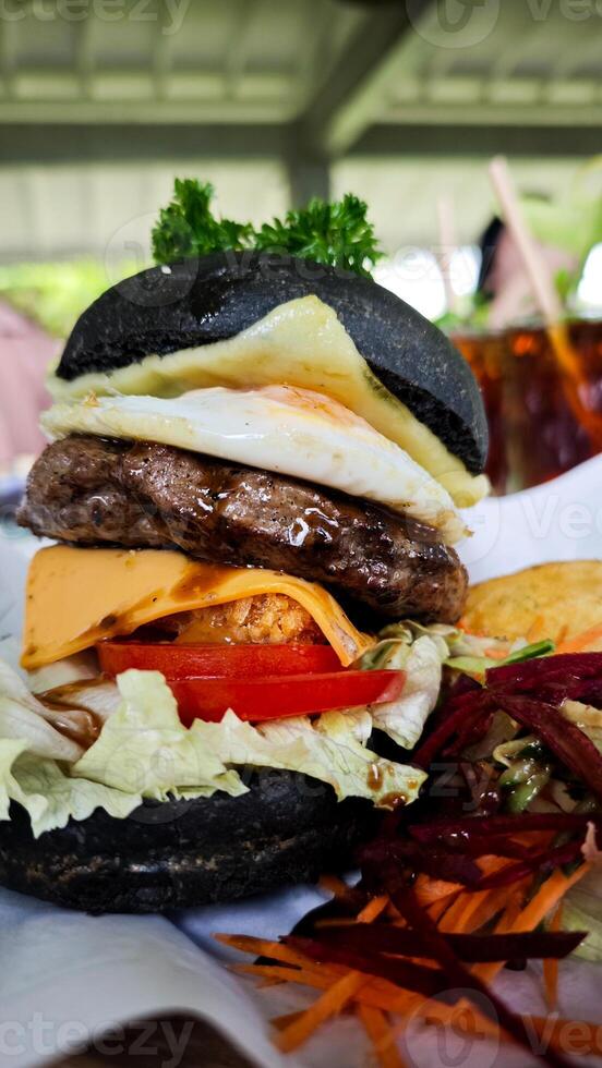 Gourmet black charcoal burger with egg, cheese, and vegetables, served with crispy wedges and fresh salad on parchment, creating a vibrant and appetizing presentation. photo