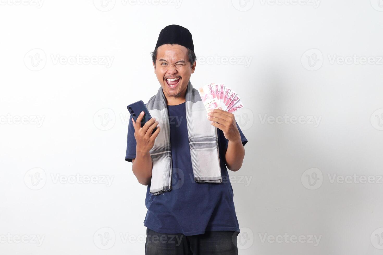Portrait of excited Asian muslim man wearing sarung scarf holding rupiah money banknote while holding and looking at his mobile phone. Isolated image on white background photo