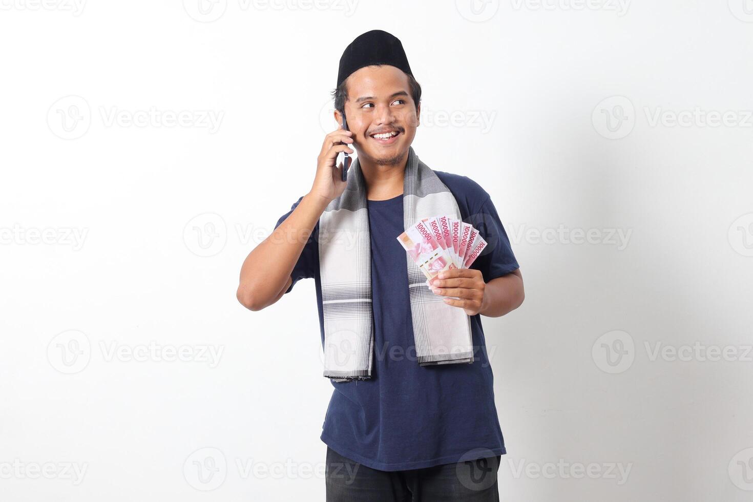 Portrait of excited Asian muslim man wearing sarung scarf holding one hundred thousand rupiah while having a conversation on his mobile phone. Isolated image on white background photo