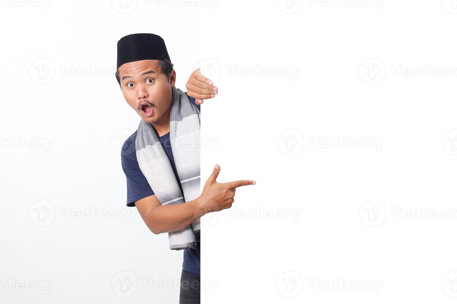 Portrait of excited Asian muslim man wearing sarung scarf hiding behind empty white billboard with mockup template and copy space for graphic. Advertising poster. Isolated on white background photo