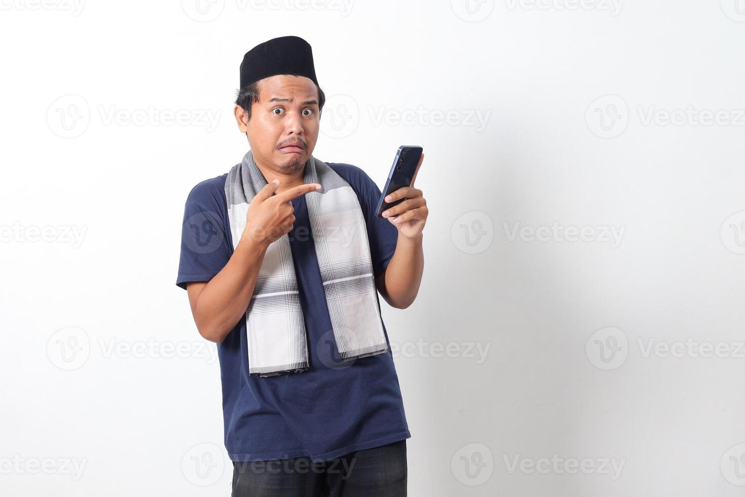 Portrait of upset Asian muslim man wearing sarung scarf and skullcap, holding mobile phone and showing shocked expression. Isolated on white background photo