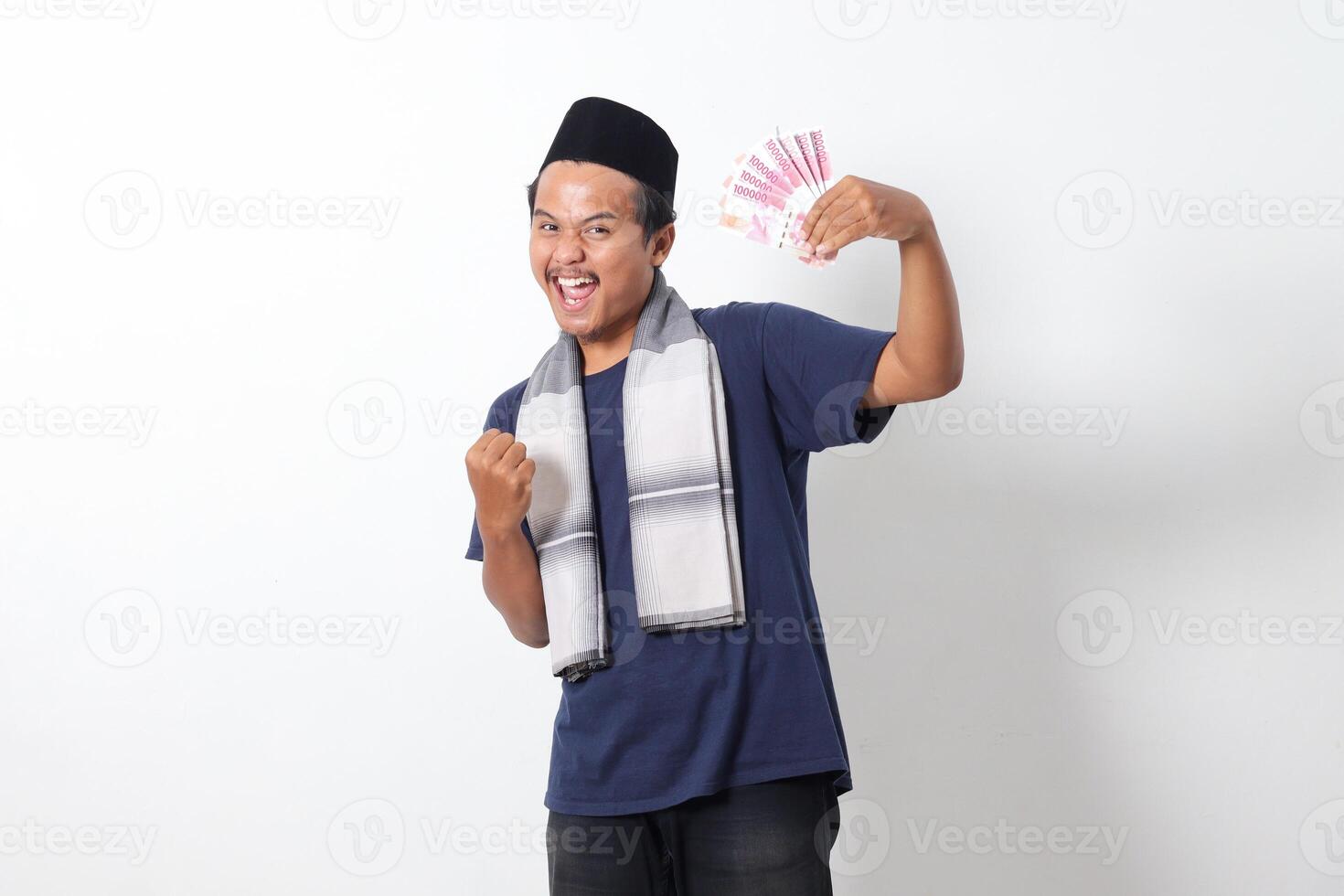 Portrait of excited Asian muslim man wearing sarung scarf holding rupiah money banknote while raising his fist, celebrating success. Financial concept. Isolated image on white background photo
