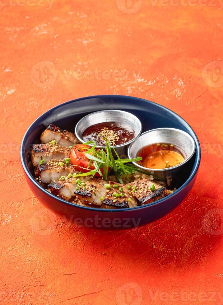 Korean Grilled Pork Belly Rice Bowl with dipping sauce with herbs isolated on orange background side view of fast food meal photo