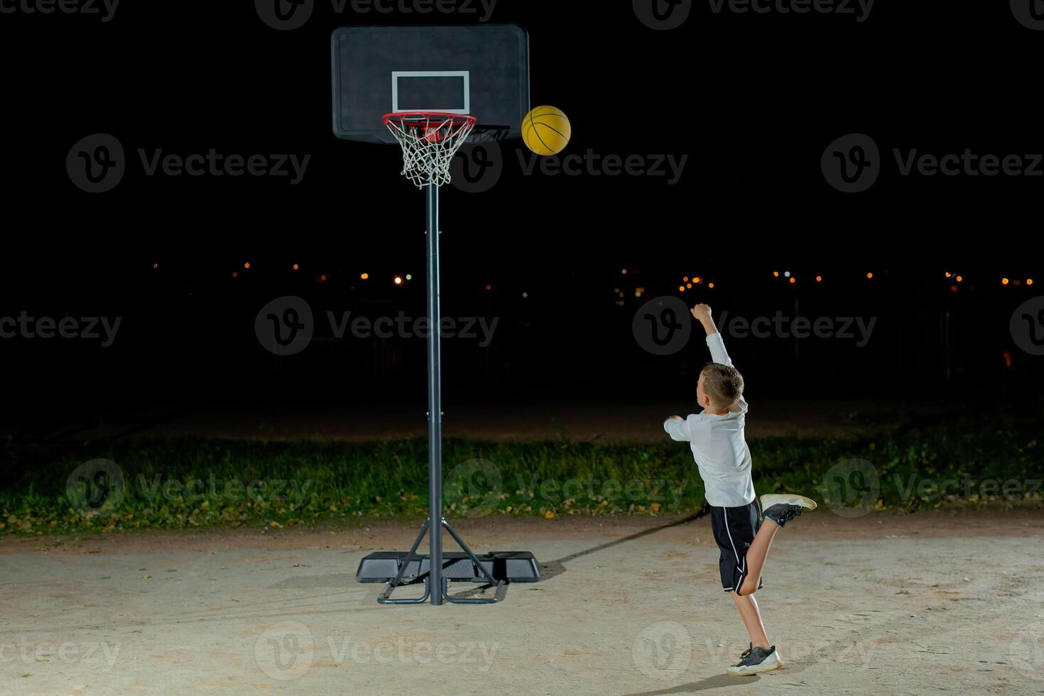 A child shoots a basketball at a hoop during nighttime practice in an outdoor playground with city lights in the background photo