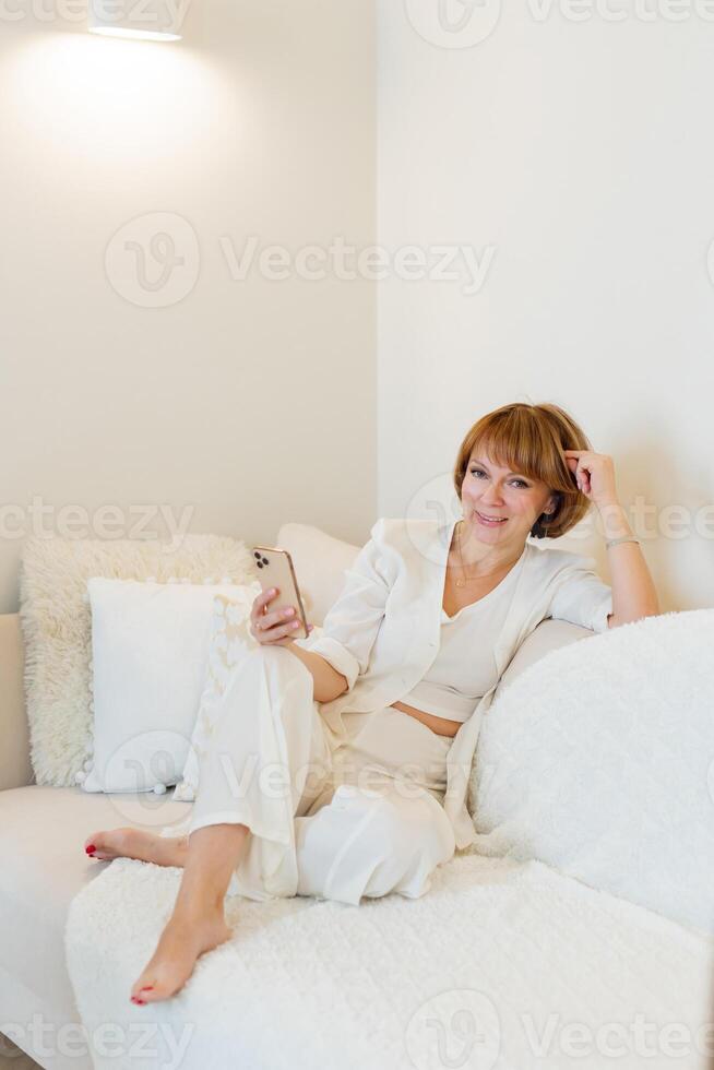 Adult beautiful woman relax siting on sofa at home and using cellphone texting chatting with friend. Happy young woman on couch holding smartphone in her hands, shoping online or check social media. photo