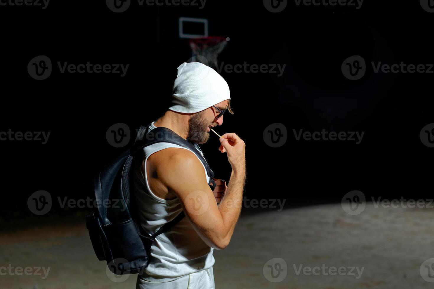 A muscular man wearing sunglasses and a beanie while holding backpack stands confidently in the dark photo