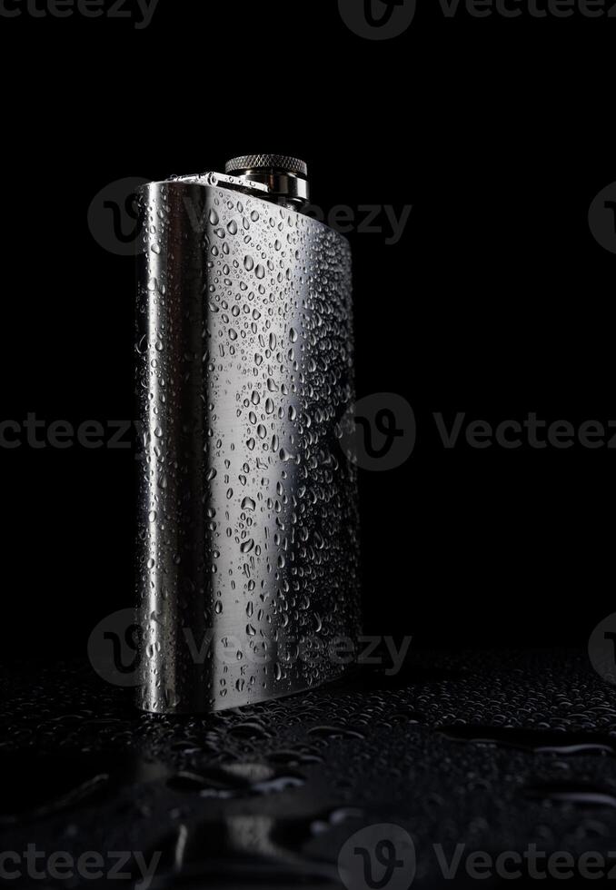 Stainless steel flask on a black background with water drops.Personal flat flask. photo