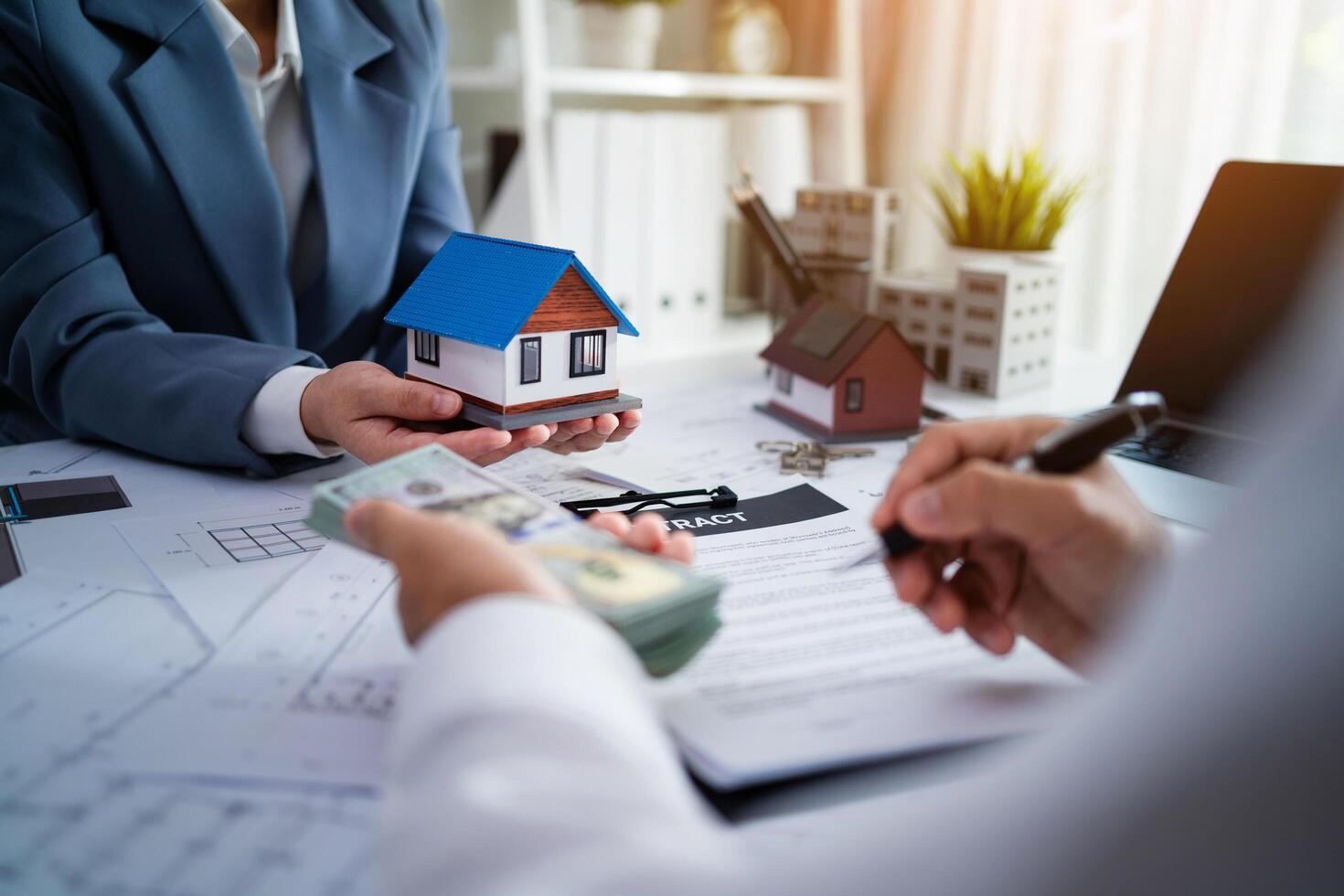 A man and a woman are sitting at a table with a house model and a stack of cash. The woman is holding the house model and the man is writing something on a piece of paper photo