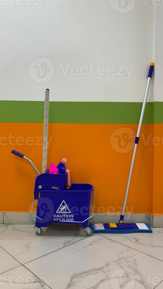 Blue trolley cleaning bucket with mop and cleaning products in corridor, vertical photography of floor cleaning service concept, cleaning bucket on wheels with inscription caution wet floor photo