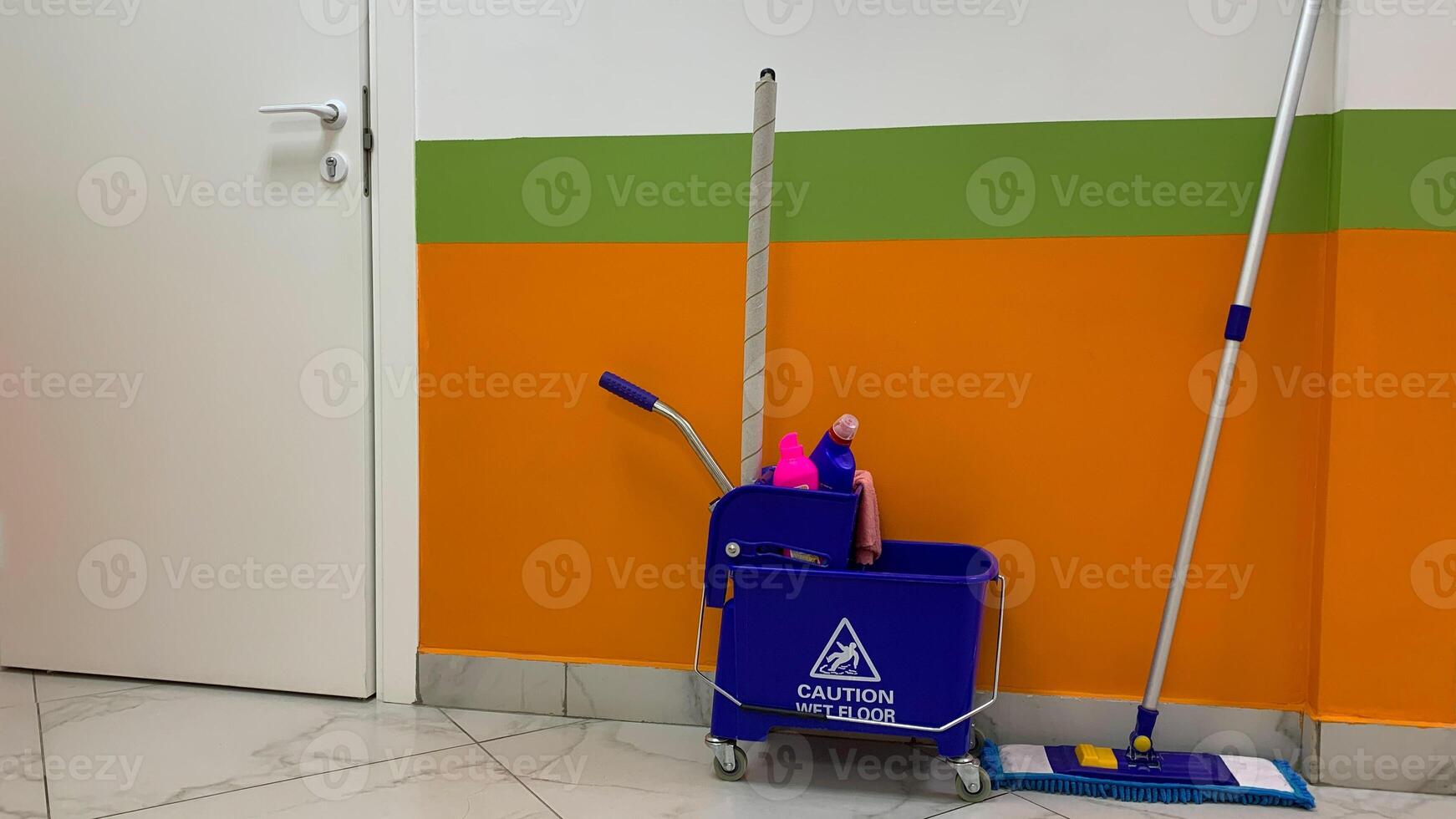 Bucket trolley with detergents and mop for cleaning floor against background of wall and door in corridor, cleanliness in public place, cart on wheels for cleaning service employee, caution wet floor photo