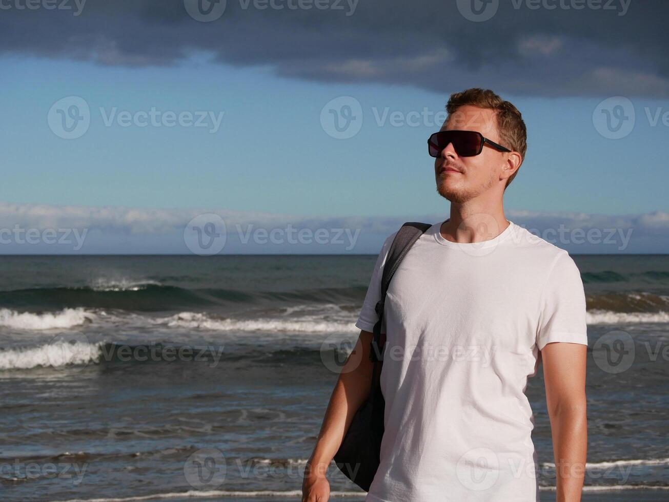 A young man with a backpack, wearing a white T-shirt and sunglasses standing on the seashore at the background of gloomy cloudy sky, with copy space. Tourism, traveling and resort recreation concept photo