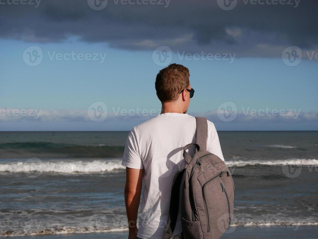 A young man with a backpack, wearing a white T-shirt and sunglasses looking at the sea at the background of gloomy cloudy sky, with copy space. Tourism, traveling and resort recreation concept photo