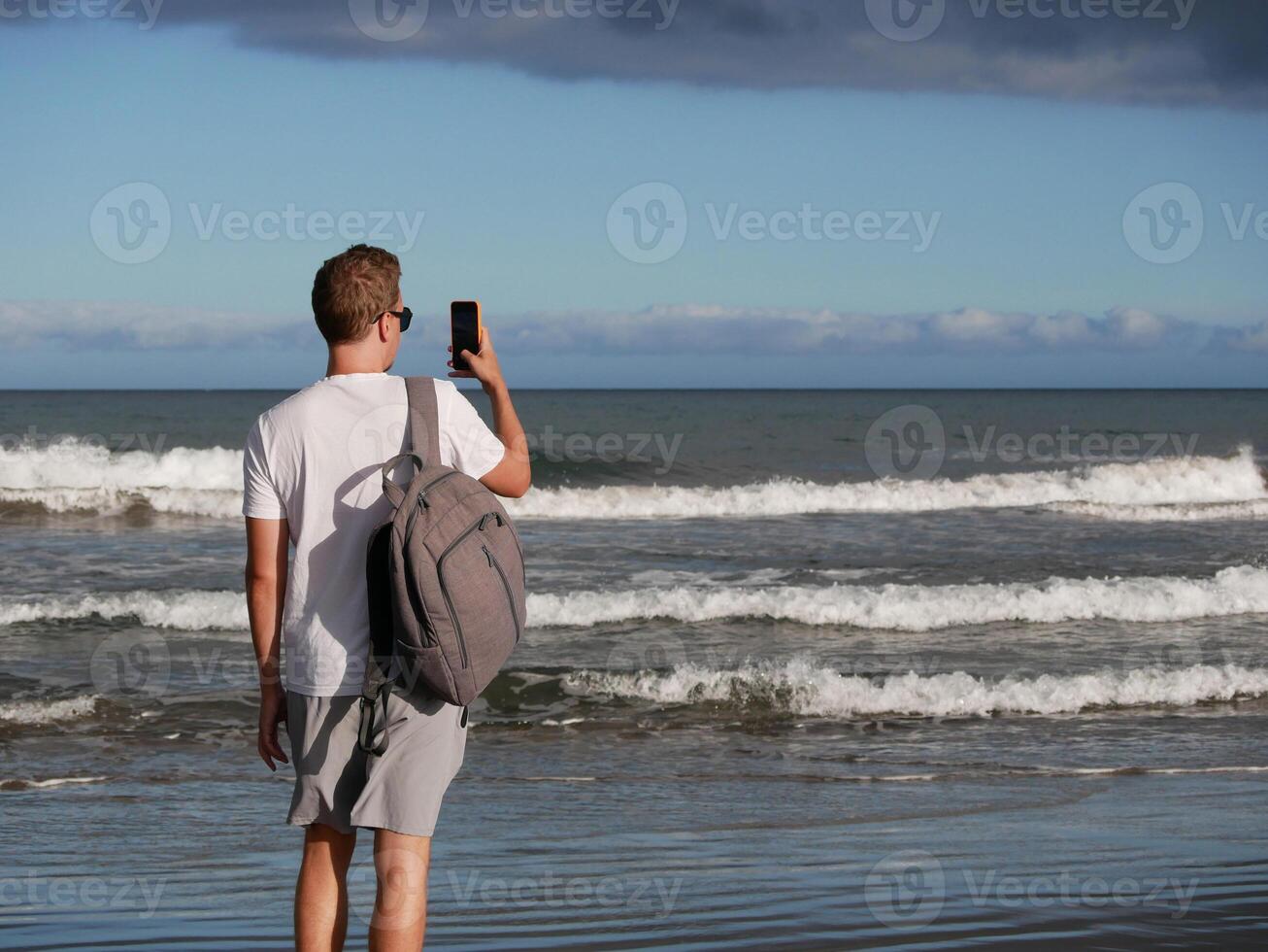 A young man with a backpack, wearing a white T-shirt and sunglasses taking a photo the sea at the background of gloomy cloudy sky, with copy space. Tourism, traveling and resort recreation concept